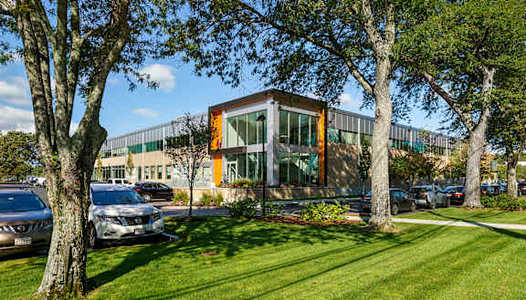 A modern building with orange and green accents is surrounded by trees and parked cars.