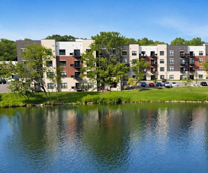 Lakeside view of Panton Mill Apartments with modern buildings, green trees, and water on a sunny day in Illinois