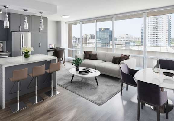 Image of a staged suite, the view is from the kitchen looking over the coffee table back to the dining room. The furniture is a contemporary adaptation of the mid-century modern style.  In this image, the TV mounted on the wall is visible.