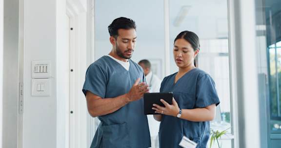 Two medical professionals in blue scrubs are discussing something on a tablet.