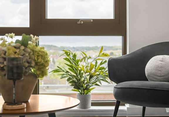 A grey chair with a white pillow sits in front of a window with a view of the outdoors.