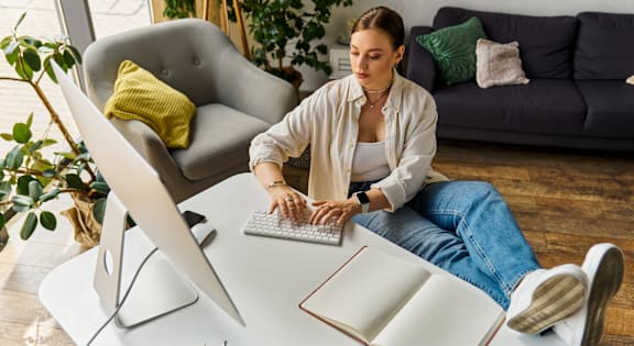A woman is sitting in front of a desk with a computer and a notebook.