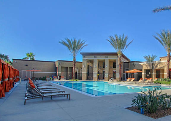 Dog-Friendly Apartments in Redlands, CA - Circa 2020 - Swimming Pool Lined with Chaise Lounges, Umbrellas, Private Cabanas with Drapery, Palm Trees Landscape Area, and Stucco-Exterior Building View.