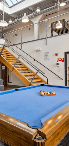A pool table is in the middle of a room with a staircase in the background.