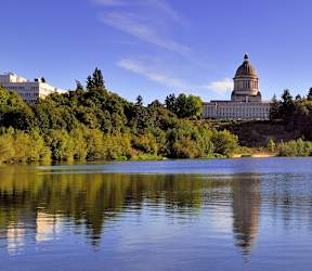 A large white building with a dome is situated on a hill with trees and is reflected in the water.