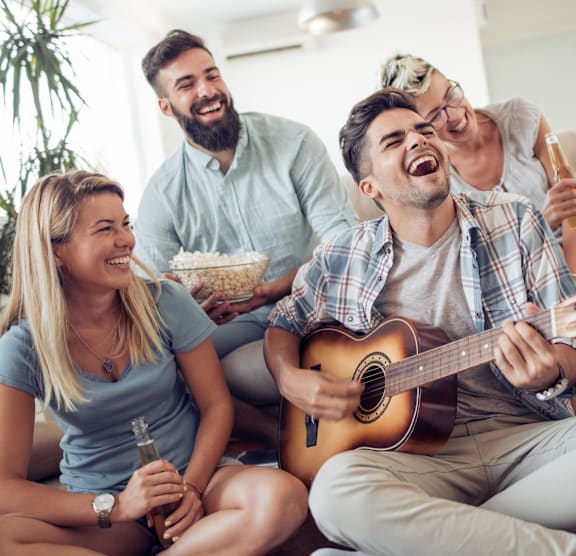 Four people are sitting on a couch, with one playing a guitar and another holding a bottle.
