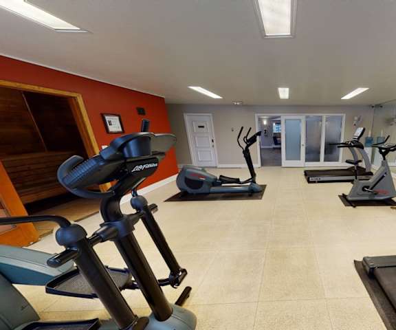 A spacious gym with cardio machines, windows, TVs, and weights with a red accent wall at the Club at Eagle Point Apartment Homes. 