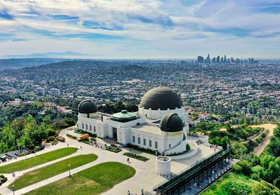 A large white building with two large domes sits in the middle of a green field with a city in the background.