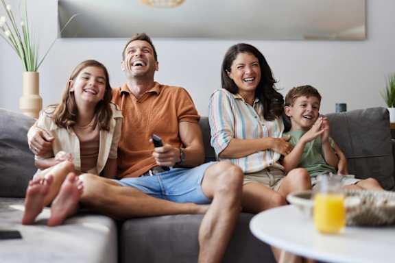 A family of four is sitting on a grey couch, watching something together.