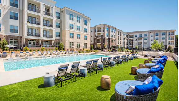 A large swimming pool surrounded by lounge chairs and umbrellas in front of apartment buildings.
