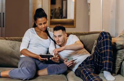 A man and a woman are sitting on a couch, both looking at a tablet.