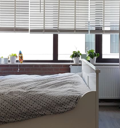 A bedroom with a bed, a window with blinds, a radiator, a bookshelf, and a small table with a plant and a vase on it.