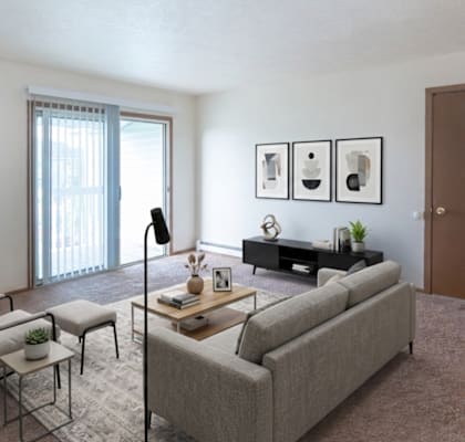 A living room with a grey couch, a chair, a small table with a vase and a plant on it, a bookshelf with a plant on top, and pictures on the wall.