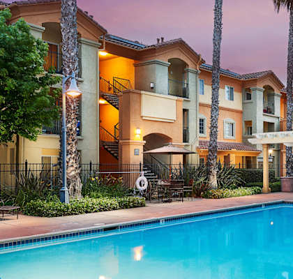 A swimming pool is surrounded by a patio with chairs and a building in the background.