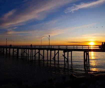 A pier with a sunset in the background and a few birds flying.
