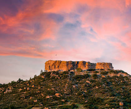 A large rock formation rises above a hill covered in greenery under a pink and orange sky.