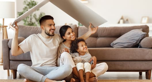 A family of three is sitting on the floor in a living room, with a couch and a lamp in the background.