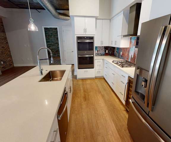 A large, modern kitchen with stainless steel appliances, kitchen utensils, white countertops with lots of room, a kitchen island with barstools and two places set for a meal, wall art, and large white cabinets at Roosevelt Row Apartment Homes. 