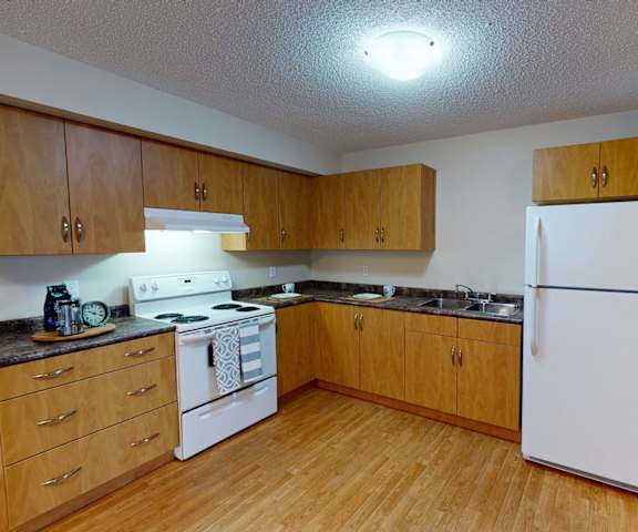 A spacious kitchen with white appliances, kitchen utensils, overhead lighting, black countertops with lots of room, a bar counter with barstools, and large medium wooden cabinets at Sonora Apartment Homes. 