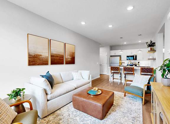 A living room with a white couch, a brown ottoman, and a TV in the background.
