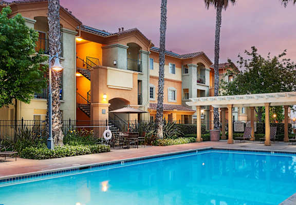 A swimming pool is surrounded by a patio with chairs and a building in the background.