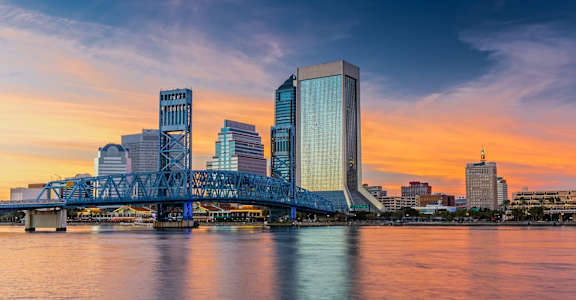 A city skyline at sunset with a bridge crossing a body of water.