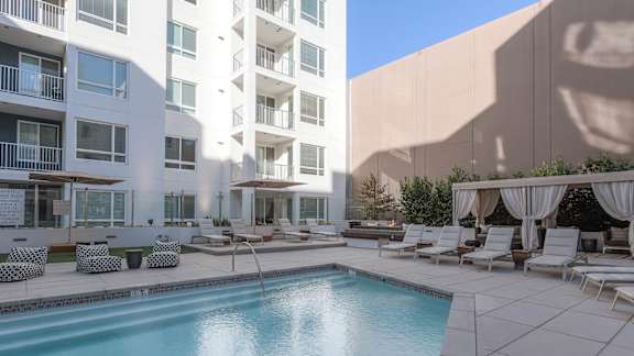 A swimming pool is surrounded by lounge chairs and umbrellas in a sunny courtyard.
