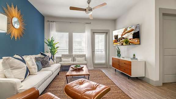 A living room with a white couch, brown leather ottoman, and a flat screen TV mounted on the wall.