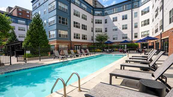 A swimming pool surrounded by lounge chairs and umbrellas in front of a multi-story building.