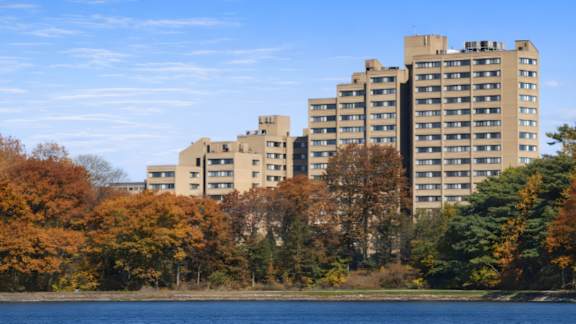 A row of apartment buildings are in the background of a lake and trees.