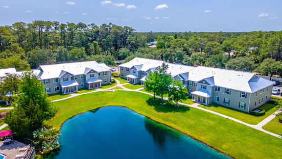 A large white building with a grey roof is surrounded by a green lawn and a blue pond.