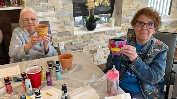 Two women holding painted pots in front of a table with paint bottles.