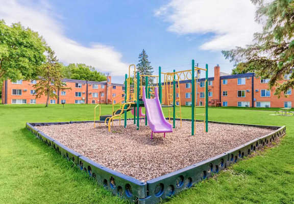 A playground with a slide, swings, and a seesaw in front of a building.