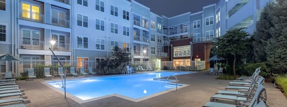 an outdoor swimming pool with chaise lounge chairs at night in front of an apartment building
