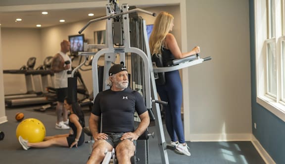 A man is sitting on a gym machine while a woman is working out on a treadmill in the background.