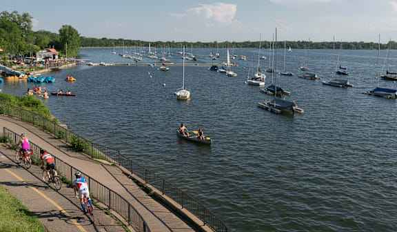 A group of people riding bikes on a path next to a body of water with boats in the background.