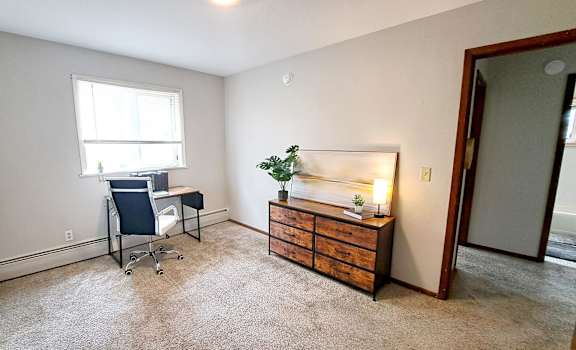 A room with a desk, chair, and a wooden cabinet with a lamp on it.