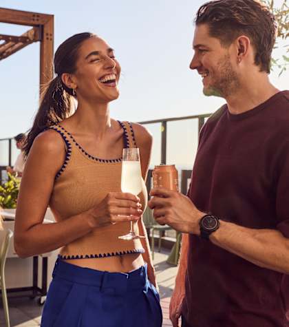 A woman in a tan top and blue pants is holding a drink and laughing with a man in a maroon shirt.