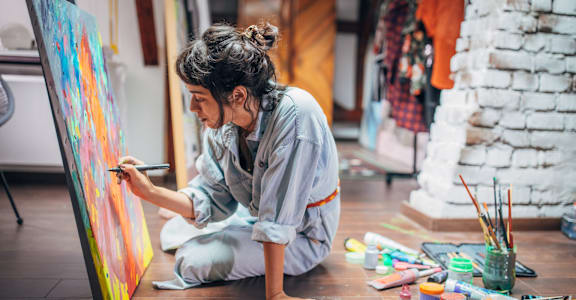 a woman sitting on the floor painting a canvas with paint brushes
