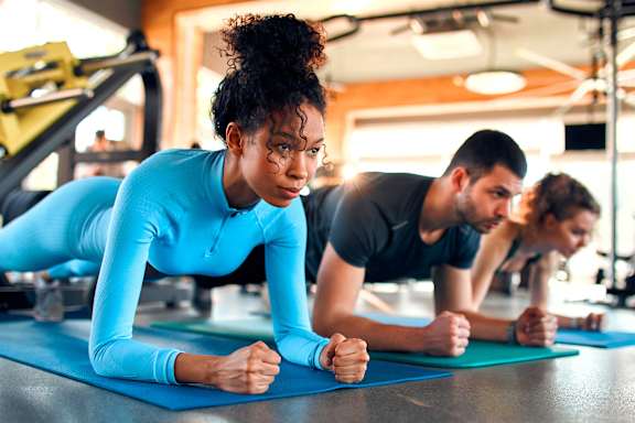 A woman in a blue top and black pants is doing push-ups on a blue mat in a gym.