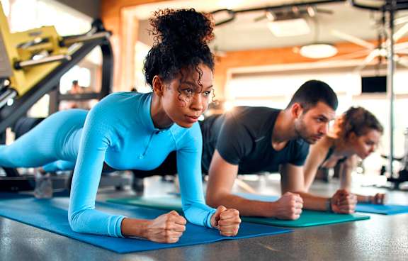 A woman in a blue top and black pants is doing push-ups on a blue mat in a gym.