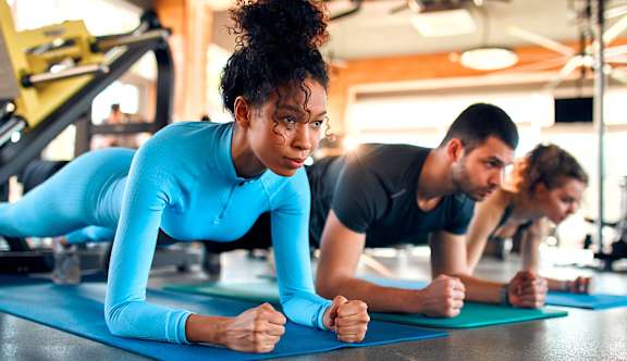 A woman in a blue top and black pants is doing push-ups on a blue mat in a gym.