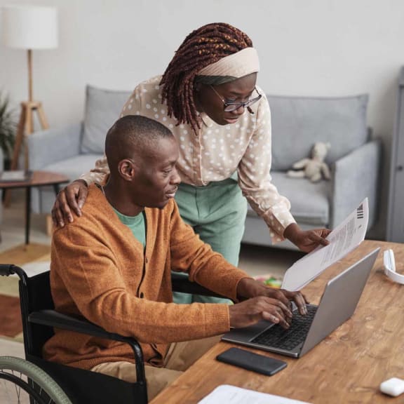 a man in a wheelchair using a laptop computer with a woman standing next to him