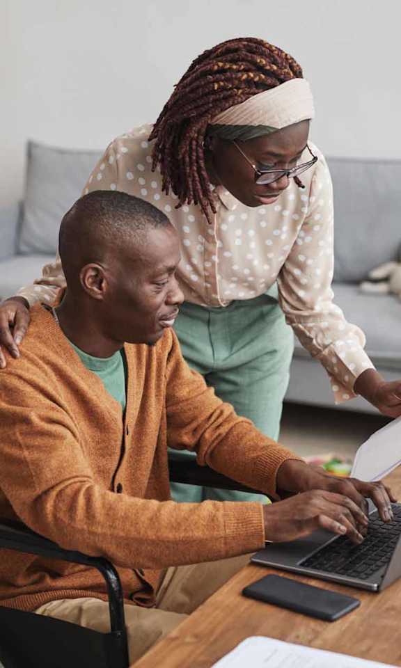 A person in a wheelchair is using a laptop with another person standing beside them.