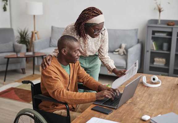 a man in a wheelchair using a laptop computer with a woman standing next to him