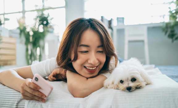 A woman is lying on a bed with a white dog, looking at a pink phone.