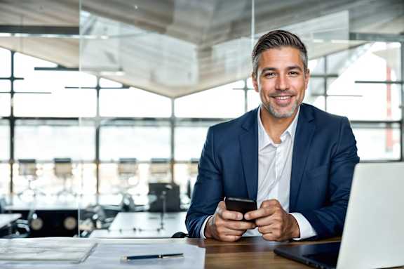 A man in a suit is sitting at a desk with a laptop and a phone in his hands.
