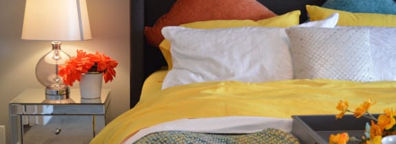 a bed with yellow sheets and pillows and a table with a lamp and flowers