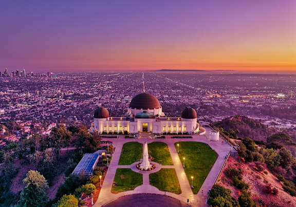 A large white building with a dome and a circular monument in front of it is surrounded by a green lawn.
