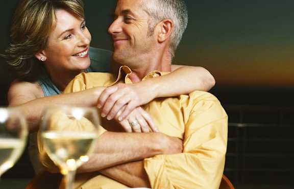 A couple in a yellow shirt are sitting close together with a glass of wine in front of them.